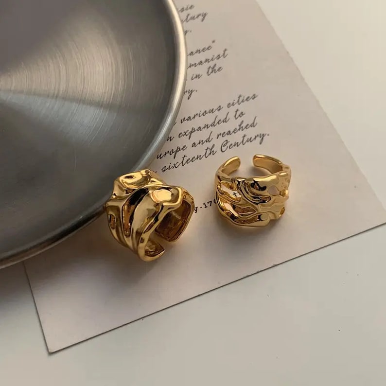 K&ouml;nnte beinhalten: Zwei goldfarbene Ringe mit abstraktem, strukturiertem Design. Die Ringe sind aus Metall und haben einen klobigen, modernen Look.
