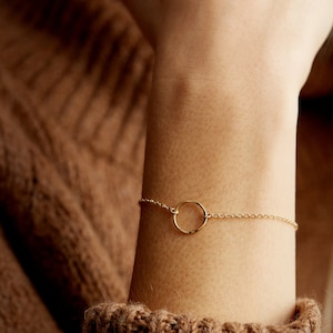 May include: A delicate gold chain bracelet with a simple circle pendant. The bracelet is shown on a person's wrist.