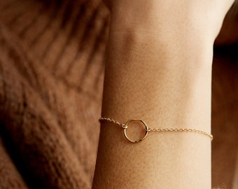 Peut inclure: Un bracelet délicat en chaîne dorée avec un pendentif rond simple. Le bracelet est présenté sur le poignet d'une personne.