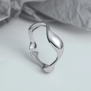 May include: A silver ring with a unique, abstract, wavy design. The ring is made of smooth metal and has a modern, minimalist style.