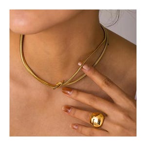 Könnte beinhalten: Eine goldfarbene Halskette mit Schlangenketten-Design und Knotendetail. Ein passender goldfarbener Ring mit quadratischem, abgerundetem Oberteil ist ebenfalls sichtbar. Der Schmuck wird auf der Haut einer Person getragen.