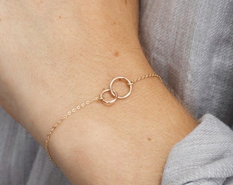 Peut inclure: Un bracelet délicat en chaîne dorée avec deux cercles entrelacés. Le bracelet est porté au poignet.