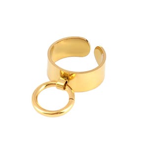 May include: A gold-colored adjustable ring with a small, dangling hoop. The ring has a thick band and an open design. The hoop is attached to the main ring with a small connector. The ring is set against a white background.