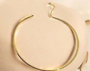 May include: A gold-toned choker necklace with a simple, minimalist design. The necklace is made of a single, continuous piece of metal that forms a circle around the neck.