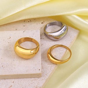 May include: Three gold and silver rings with a rounded, smooth design. The rings are on a light beige surface.