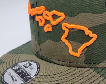 Può includere: Un cappellino da baseball mimetico con la scritta "Hawaii" ricamata in arancione brillante. Il cappellino ha una visiera piatta e una chiusura a scatto. Il logo New Era 9FIFTY è visibile sulla visiera.