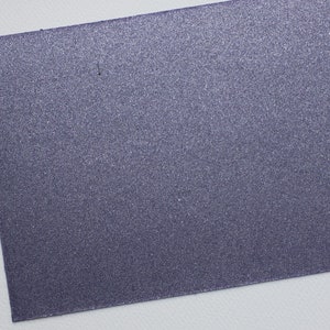 Könnte beinhalten: Ein rechteckiges Blatt dunkelblaues Papier mit einer glitzernden, strukturierten Oberfläche. Das Papier liegt vor einem weißen Hintergrund, wobei das Blatt angewinkelt ist, um seine gesamte Oberfläche zu zeigen.