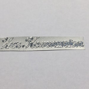 Könnte beinhalten: Ein rechteckiges, silberfarbenes Metallschild mit dem geprägten Text "Zur Kommunion" in einer dekorativen Schrift. Das Schild ist leicht gebogen und liegt vor einem schlichten, hellen Hintergrund.