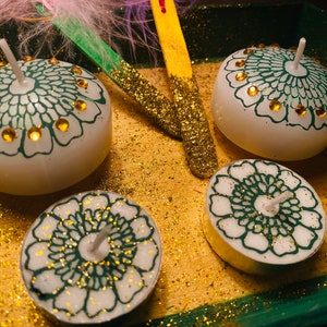 Könnte beinhalten: Vier weiße Kerzen mit grünen und goldenen Designs. Die Kerzen stehen auf einer goldenen Glitzerfläche.