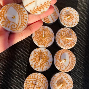 Puede incluir: Una colección de diez velas de té blancas con intrincados diseños de henna dorados. Las velas están dispuestas en círculo sobre una superficie negra.