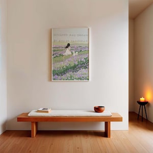 May include: A white framed print featuring a colourful illustration of a girl and a dog in a field of purple flowers. The text on the print reads "Designed and Drawn by Ashley Yamakawa". A wooden bench with a light brown cushion sits in front of the print.