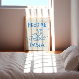 以下が含まれることがあります： ベージュの額縁に入ったプリントで、青い文字で「Feed Me Pasta」と書かれています。テキストの上に、フォークを持った手を青い線で描いた絵があり、フォークの先にはパスタの糸が乗っています。パスタはボウルに入っています。