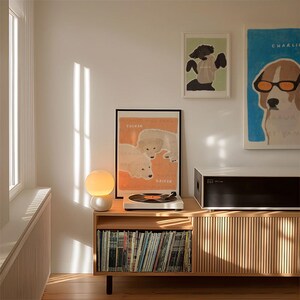 May include: A wooden record player stand with a record player on top, a lamp, and a shelf filled with vinyl records. There are three framed prints on the wall behind the stand, two of which feature dogs.