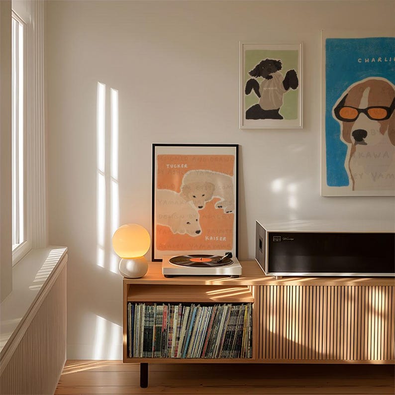 May include: A wooden record player stand with a record player on top, a lamp, and a shelf filled with vinyl records. There are three framed prints on the wall behind the stand, two of which feature dogs.