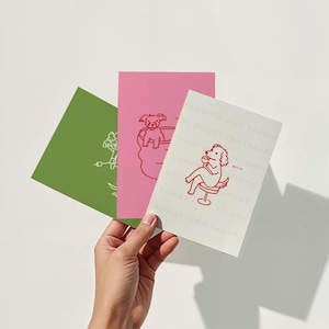 May include: Three colorful art prints held by a hand. The prints are green, pink, and off-white. Each print features a red line drawing of a dog in various poses. The off-white print has text that reads "DESIGNED AND DRAWN BY ASHLEY YAMAKAWA".