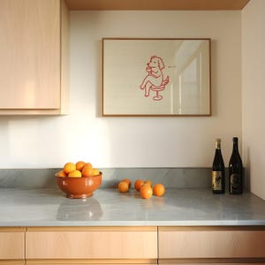 May include: A kitchen scene featuring a framed artwork with a red illustration of a dog-like figure seated on a stool, holding a cup. A terracotta bowl filled with oranges sits on a countertop, with additional oranges scattered nearby. Two wine bottles are also on the counter.