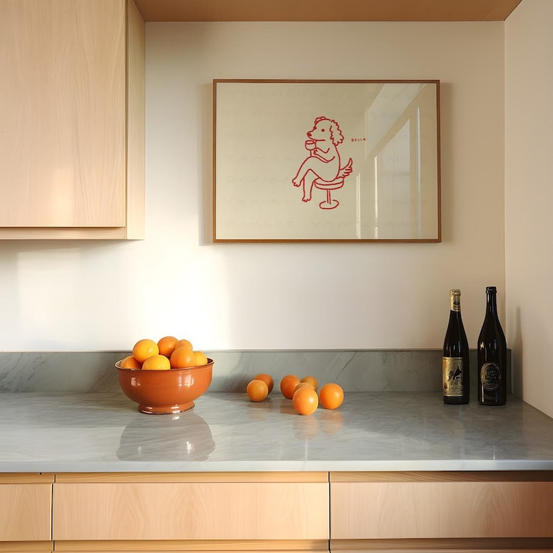 May include: A kitchen scene featuring a framed artwork with a red illustration of a dog-like figure seated on a stool, holding a cup. A terracotta bowl filled with oranges sits on a countertop, with additional oranges scattered nearby. Two wine bottles are also on the counter.