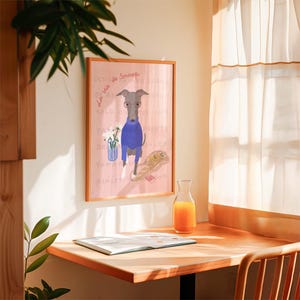 May include: A framed illustration of a grey dog wearing a blue jumper and glasses. The dog is standing on a pink background with text and a basket of flowers. The frame is brown wood.
