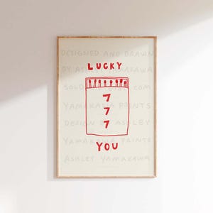 Peut inclure: Une impression encadr&eacute;e avec une illustration rouge d'une bo&icirc;te d'allumettes avec le mot "LUCKY" au-dessus et le nombre "7" r&eacute;p&eacute;t&eacute; quatre fois &agrave; l'int&eacute;rieur de la bo&icirc;te d'allumettes. Le texte "DESIGNED AND DRAWN BY ASHLEY YAMAKAWA" est au-dessus de la bo&icirc;te d'allumettes et le texte "YOU" est en dessous de la bo&icirc;te d'allumettes. Le texte "YAMAKAWA PRINTS" est en dessous du mot "YOU".