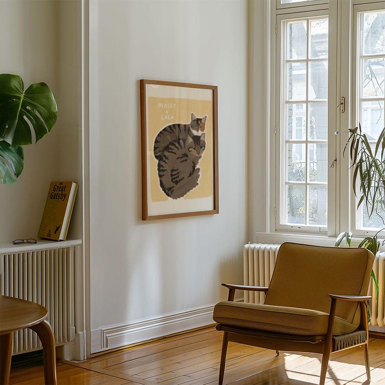May include: A framed print of two cats, one on top of the other, with the text "Maisy & Lila" in a light yellow font. The print is on a white background and is framed in a light brown wood frame. The print is hanging on a white wall in a room with a brown wooden floor.