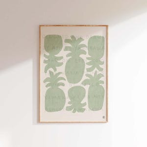 Peut inclure: Une impression encadrée avec un fond vert clair représentant un motif d'ananas stylisés. L'impression porte le texte "Designed and Drawn by Ashley Yamakawa" en brun clair.