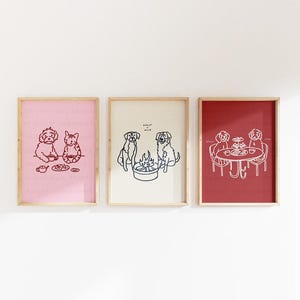 Może przedstawiać: Trzy oprawione grafiki z rysunkami zwierząt. Pierwsza grafika ma różowe tło z kotem i psem. Druga grafika ma kremowe tło z psami i ogniskiem. Trzecia grafika ma czerwone tło z psami przy stole.