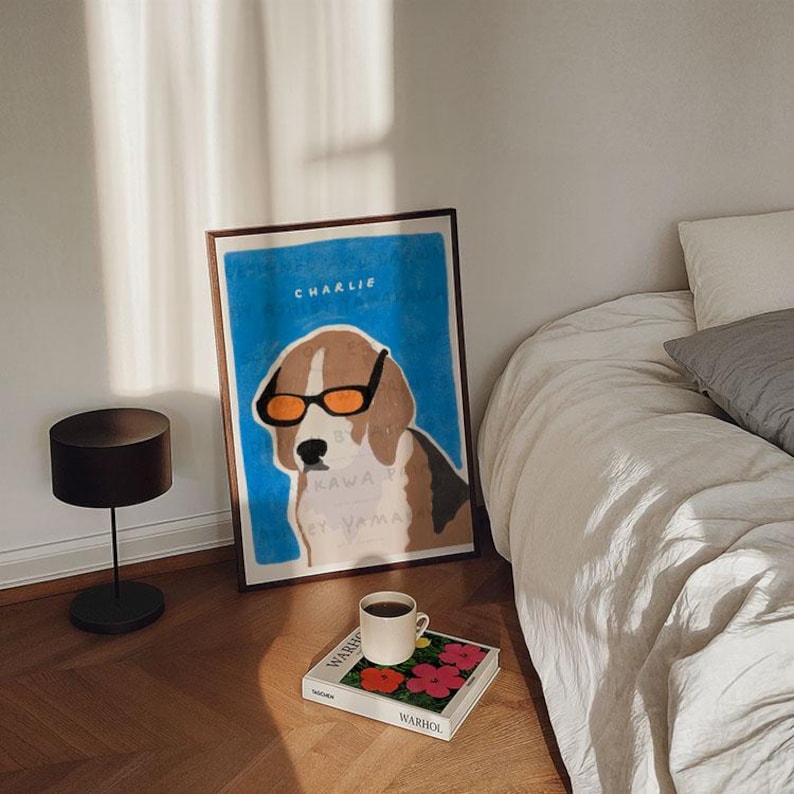 May include: A framed illustration of a dog wearing sunglasses with the text "Charlie" and "Kawaii Eyewear" on a blue background. The frame is sitting on a table next to a book and a cup of coffee.