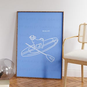 May include: A framed art print featuring a white line drawing of a dog on a paddleboard with a paddle, set against a blue background. The word "HENLEY" is above the dog. The frame is brown.