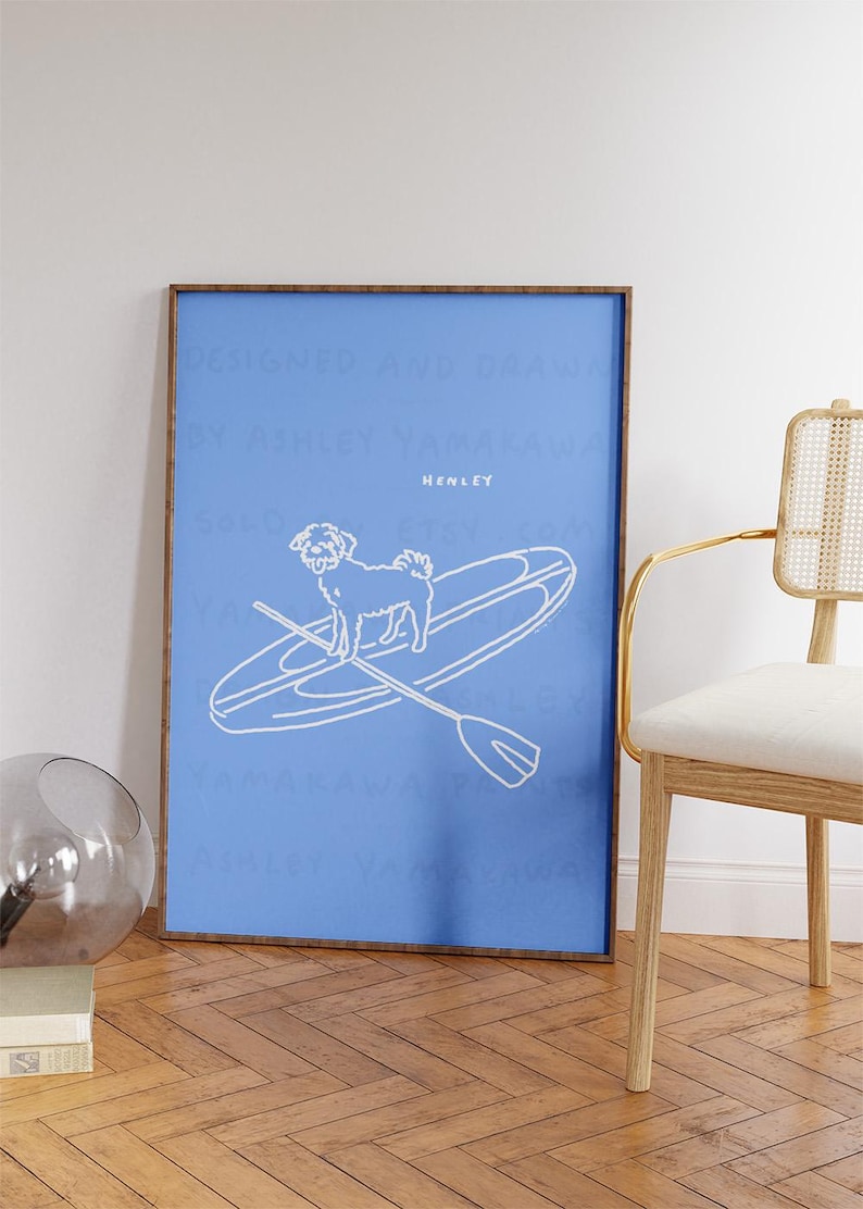 May include: A framed art print featuring a white line drawing of a dog on a paddleboard with a paddle, set against a blue background. The word "HENLEY" is above the dog. The frame is brown.