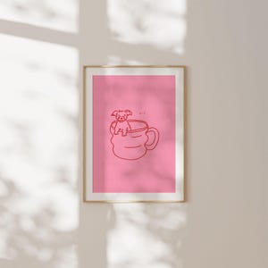 May include: A framed print featuring a pink background with a red line drawing of a dog sitting in a coffee mug. The artwork has a minimalist style and is displayed in a gold frame.