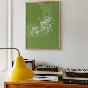 May include: A framed print with a green background features a white line drawing of a dog roasting a marshmallow over a campfire. The print is in a wooden frame and hangs above a wooden cabinet with a yellow lamp, audio equipment, and a keyboard.
