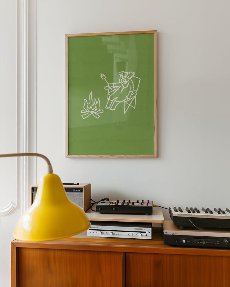 May include: A framed print with a green background features a white line drawing of a dog roasting a marshmallow over a campfire. The print is in a wooden frame and hangs above a wooden cabinet with a yellow lamp, audio equipment, and a keyboard.
