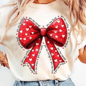 May include: Cream-colored t-shirt featuring a large red bow with white heart patterns. The bow is surrounded by text that reads "You are beautiful, loved, worthy, brave, smart, enough, capable, unique, kind, and strong."