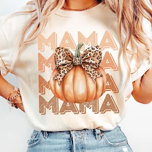 May include: A white t-shirt with the word "MAMA" repeated four times in a brown and orange gradient. A large orange pumpkin with a brown stem and a leopard print bow is centered on the shirt.