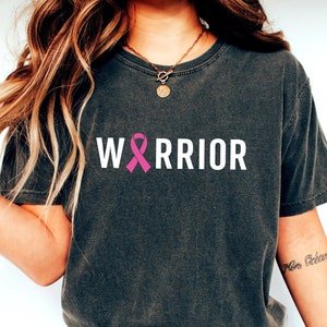 May include: Dark gray t-shirt with the word "WARRIOR" in white block letters and a pink ribbon graphic. The shirt is a crew neck style and has short sleeves. The shirt is made of a soft material.