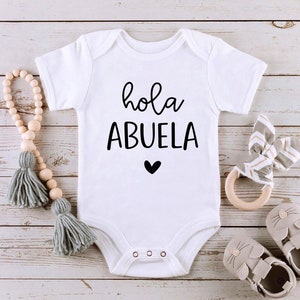 May include: White baby onesie with black text that says "hola abuela" and a small black heart. The onesie is on a wooden background with other baby items.