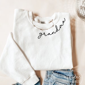 Może przedstawiać: Biała bluza z napisem "grandma" napisanym czarnym, ozdobnym pismem na przodzie. Bluza jest złożona i leży na jasnobrązowej powierzchni z parą niebieskich dżinsów.