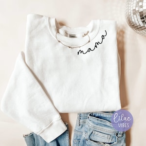 Puede incluir: Sudadera blanca con la palabra "mama" escrita en cursiva negra en la parte delantera. La sudadera está sobre una superficie rosa claro con un par de pantalones vaqueros azules y una bola de discoteca plateada.
