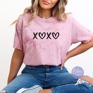 May include: A pink tie-dye t-shirt with the text "XOXO" in black. The shirt is worn by a person with long brown hair and is paired with blue jeans.