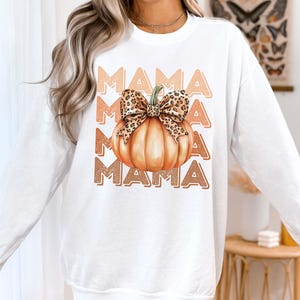 May include: White sweatshirt with a graphic of a pumpkin with a leopard print bow and the word "MAMA" repeated in a vintage style font.