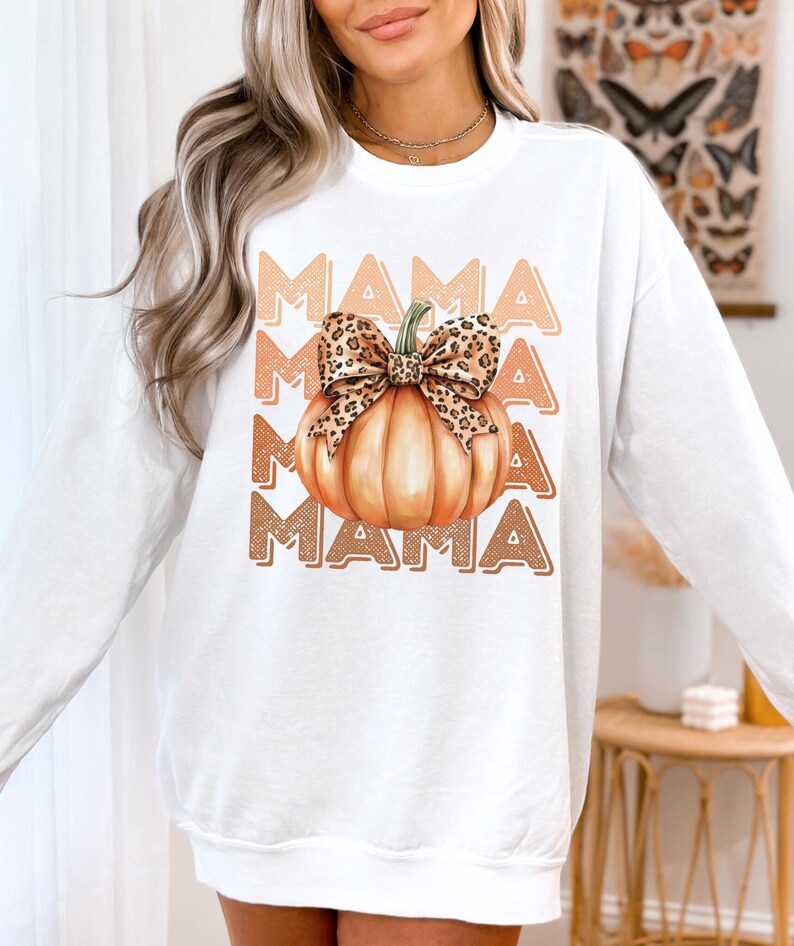 May include: White sweatshirt with a graphic of a pumpkin with a leopard print bow and the word "MAMA" repeated in a vintage style font.