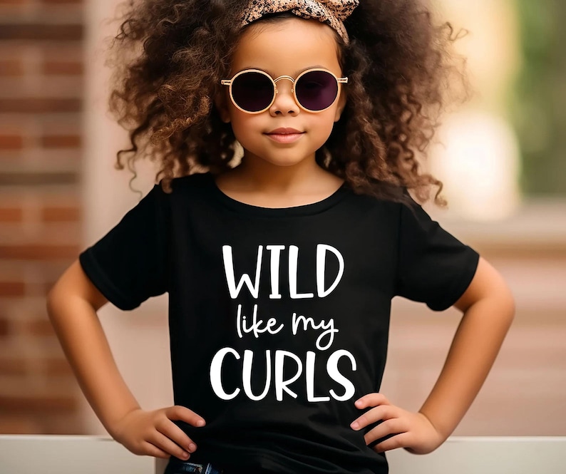May include: A young girl wearing a black t-shirt with the text "WILD like my CURLS" in white. She is wearing round sunglasses and a headband.