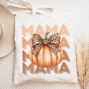 May include: A white tote bag with the text "MAMA" repeated four times in a brown and orange color scheme. A pumpkin with a leopard print bow is centered on the bag.