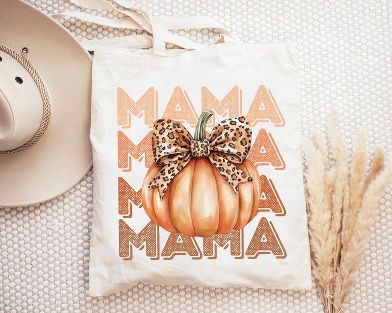 May include: A white tote bag with the text "MAMA" repeated four times in a brown and orange color scheme. A pumpkin with a leopard print bow is centered on the bag.