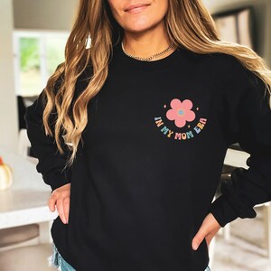 Puede incluir: Sudadera negra con una flor rosa y el texto "In My Mom Era" en colores arco&iacute;ris.