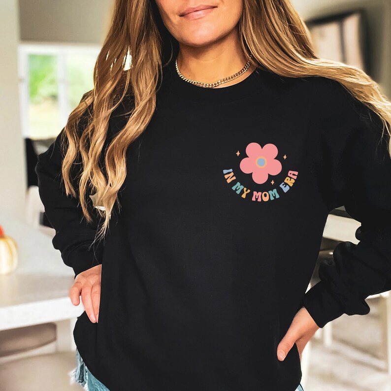 Puede incluir: Sudadera negra con una flor rosa y el texto "In My Mom Era" en colores arco&iacute;ris.