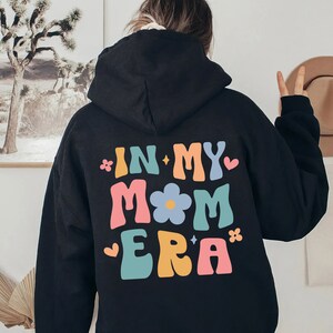 Puede incluir: Sudadera con capucha negra con el texto "In My Mom Era" en letras retro de colores con un gr&aacute;fico de flores.