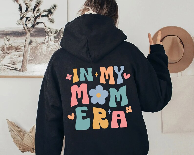 Puede incluir: Sudadera con capucha negra con el texto "In My Mom Era" en letras retro de colores con un gr&aacute;fico de flores.