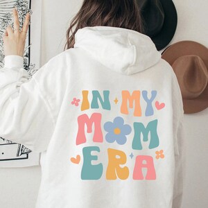 Puede incluir: Sudadera blanca con un gr&aacute;fico colorido en la espalda que dice "In My Mom Era" con una flor y estrellas.