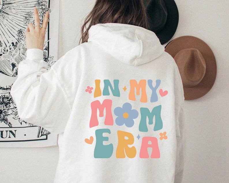 Puede incluir: Sudadera blanca con un gr&aacute;fico colorido en la espalda que dice "In My Mom Era" con una flor y estrellas.